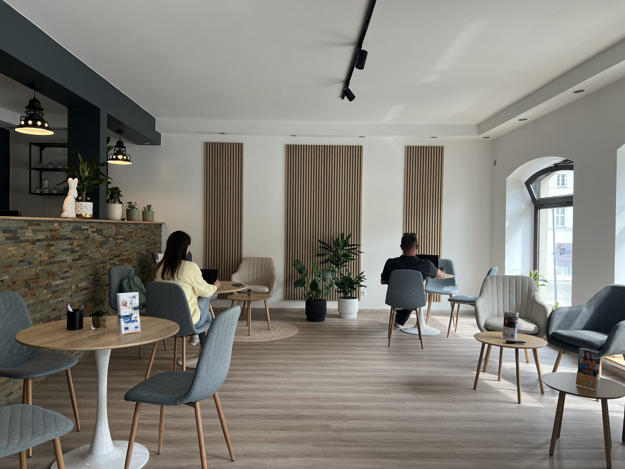 DigiUp Strausberg - Ein digitaler Ort für alle! - Modernes Raumkonzept, lichtdurchflutetes Begegnungs- und Lernraum mit stilvoller Einrichtung – Menschen sitzen an Tischen und arbeiten oder unterhalten sich.